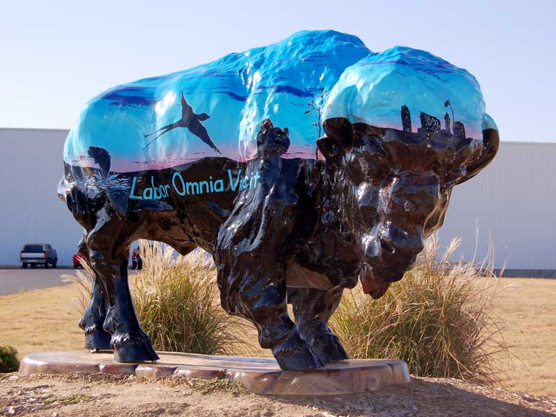 Vincit Painted buffalo sculpture with prairie horizon and sunrise. Downtown Bartlesville skyline. Local animal silhouettes dot horizon.