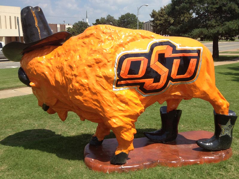 OSU Buffalo Orange painted buffalo sculpture wearing a black cowboy hat, Oklahoma State University logo on body and black cowboy boots on back legs.